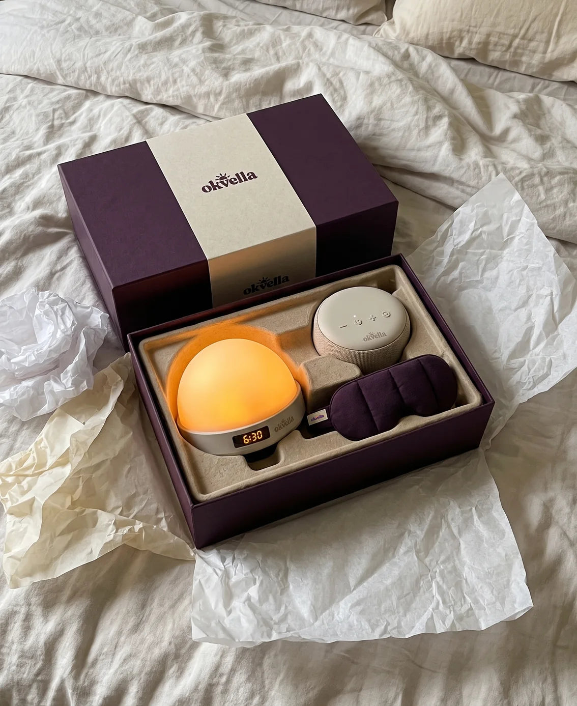 Open okvella gift box with the sunrise light, sound machine and mask on a bed